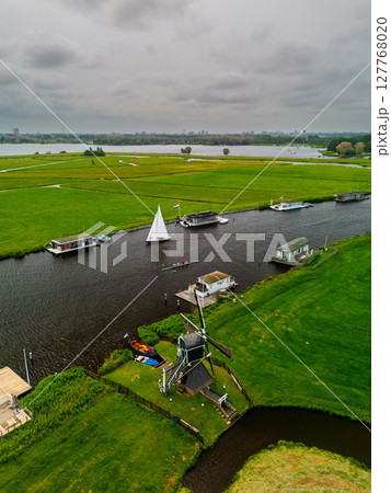 An aerial view of a Dutch countryside with a traditional windmill near a river. A small sailboat floats on the water, surrounded by green fields, farm buildings, and a cloudy sky. An aerial view of a Dutch countryside with a traditional windmill near a river. A small sailboat floats on the water, surrounded by green fields, farm buildings, and a cloudy sky. 127768020