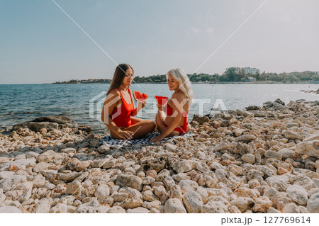 Watermelon Beach Summer: Two women enjoy refreshing watermelon slices on a rocky beach during a sunny summer day. 127769614