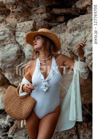 Woman Beachwear Summer Fashion Posing - A woman in a white swimsuit and a straw hat is standing in front of a rock wall. Woman Beachwear Summer Fashion Posing - A woman in a white swimsuit and a straw hat is standing in front of a rock wall. 127769708