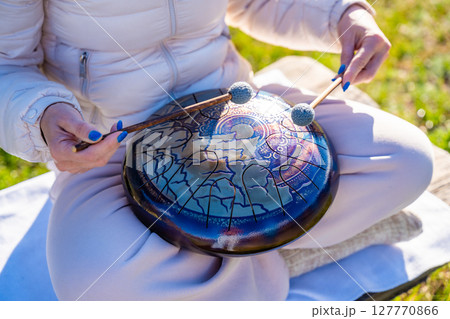 Close-up of hands playing a steel tongue drum with mallets against the backdrop of Altai mountains and river. Concept of sacred sound, touch of nature, and mindful musical ritual Close-up of hands playing a steel tongue drum with mallets against the backdrop of Altai mountains and river. Concept of sacred sound, touch of nature, and mindful musical ritual 127770866