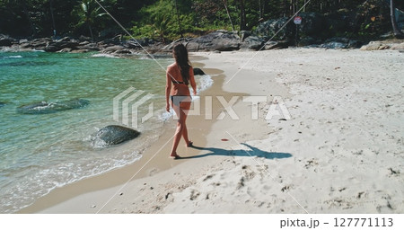 A woman strolls barefoot on a sandy beach, enjoying the warm sun and gentle waves. The clear turquoise water laps at the shore while rocky outcrops frame the scene. A woman strolls barefoot on a sandy beach, enjoying the warm sun and gentle waves. The clear turquoise water laps at the shore while rocky outcrops frame the scene. 127771113