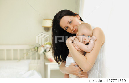 Happy young mother holding her cute baby standing together in white bedroom at home Happy young mother holding her cute baby standing together in white bedroom at home 127771850