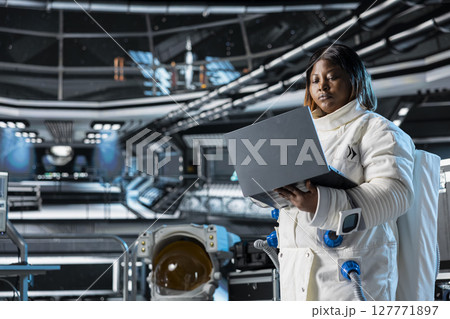 Astronaut using laptop to confirm suit integrity, monitoring life support systems. Orbital station crew member testing gear during routine equipment check using notebook, conducting safety checks 127771897