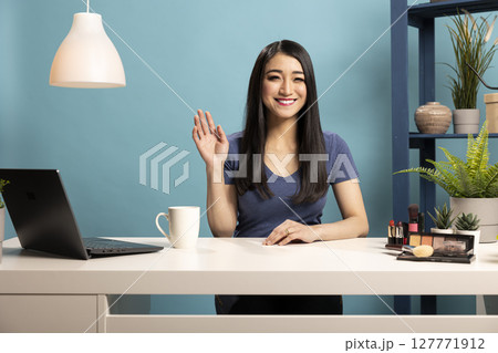 Smiling asian woman seated at desk, waving hello to her subscribers during a live broadcast. Beautiful female vlogger doing a greeting gesture with her hand towards the camera, recording a video. Smiling asian woman seated at desk, waving hello to her subscribers during a live broadcast. Beautiful female vlogger doing a greeting gesture with her hand towards the camera, recording a video. 127771912