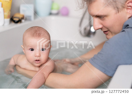 Cute baby bathing and swimming in the bath. Baby learning to swim and playing sports. Father enjoys bath time with his curious baby in a cozy bathroom. Fatherhood concept. Selective focus. 127772450