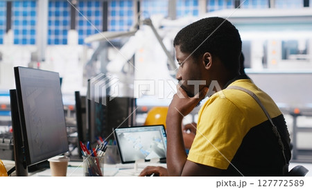 Black worker programming robotic assembly line in solar cell factory, working with high tech software for automation. Male engineer testing photovoltaic system, machinery. Camera A. 127772589