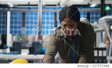 African american female expert reviewing solar panels production line, doing quality control in a photovoltaics factory. Analyzing data reports and insight to create solar power. Camera B. African american female expert reviewing solar panels production line, doing quality control in a photovoltaics factory. Analyzing data reports and insight to create solar power. Camera B. 127772590