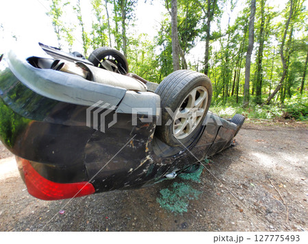 Damaged vehicle is flipped on its side along wooded dirt path, with vehicle collision 127775493