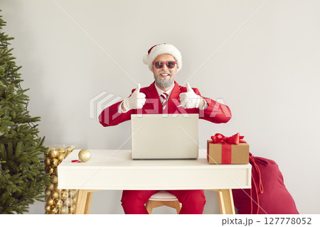 Funny happy modern Santa sitting at table with laptop in his office and giving thumbs up 127778052