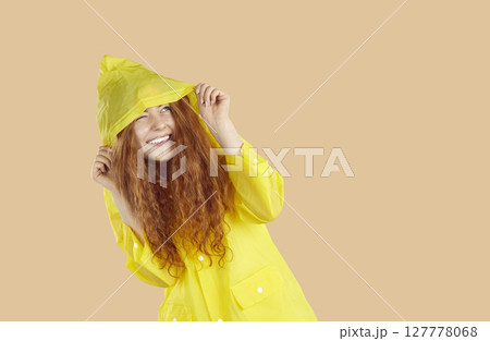 Happy young woman hiding from rain under yellow raincoat on beige copy space background 127778068