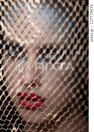 Abstract studio portrait of beautiful woman with fancy make-up looking through brown grid 127779638