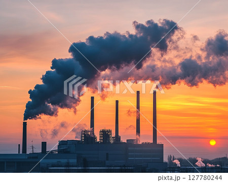 A factory with multiple smokestacks spews thick plumes of smoke against a fiery orange and red sunset sky, highlighting industrial pollution. 127780244