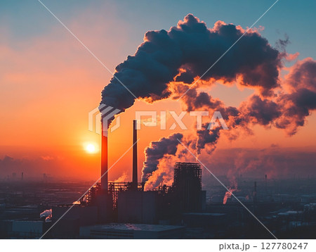 A factory with multiple smokestacks spews thick plumes of smoke against a fiery orange and red sunset sky, highlighting industrial pollution. 127780247