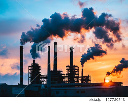 A factory with multiple smokestacks spews thick plumes of smoke against a fiery orange and red sunset sky, highlighting industrial pollution. 127780248