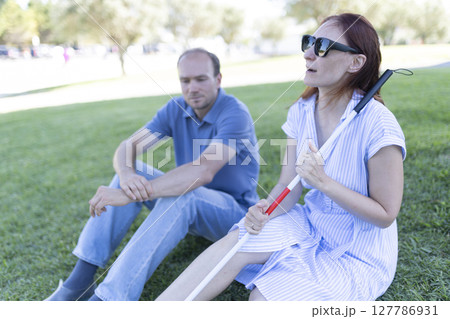 Blind woman holding white cane sitting with man in park 127786931