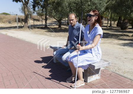 Blind woman holding white cane sitting with friend on bench in park Blind woman holding white cane sitting with friend on bench in park 127786987