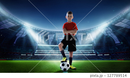 Young soccer player standing confidently on lit stadium field with ball. Youth sports ambition. Young soccer player standing confidently on lit stadium field with ball. Youth sports ambition. 127789514