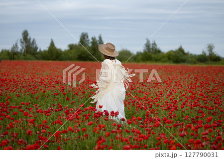 Young woman in a white dress and hat walks among summer poppies 127790031