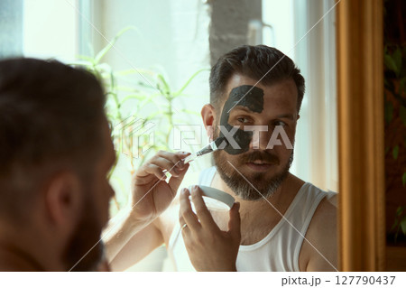 Natural beauty. Man applying face mask in mirror for skincare routine in bright bathroom 127790437