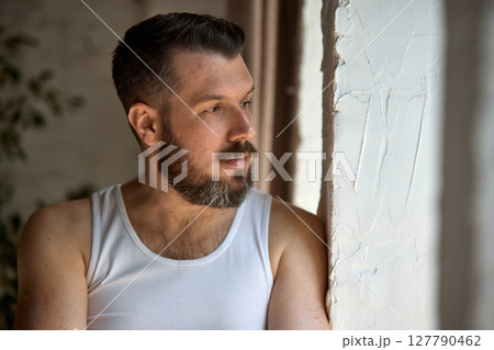 Pensive bearded man in white tank top gazing out window in soft natural light indoors 127790462