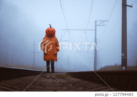 Girl in orange jacket with carved pumpkin head stands on foggy railway 127794485
