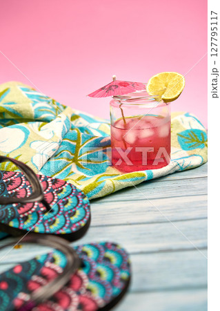 Refreshing pink drink with an umbrella and a slice of lemon on a beach towel with a tropical pattern and flip flops in the foreground with a pink background. Refreshing pink drink with an umbrella and a slice of lemon on a beach towel with a tropical pattern and flip flops in the foreground with a pink background. 127795117