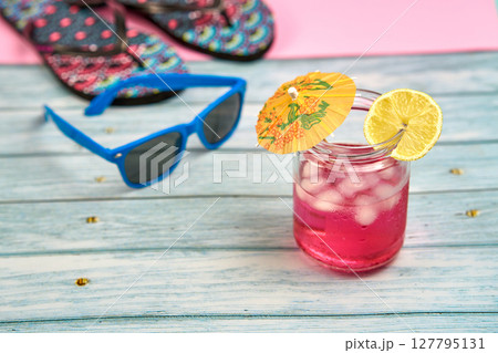 Watermelon and lemon drink with an umbrella and a slice of lemon, blue sunglasses and flip flops on a wooden table with a pink background. Watermelon and lemon drink with an umbrella and a slice of lemon, blue sunglasses and flip flops on a wooden table with a pink background. 127795131