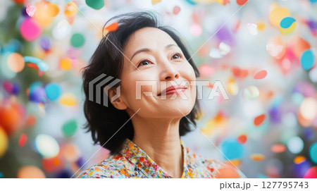 Happy mature Asian woman looks up with joyful smile as colorful confetti falls around her. Ideal for themes of celebration, success and positive emotions Happy mature Asian woman looks up with joyful smile as colorful confetti falls around her. Ideal for themes of celebration, success and positive emotions 127795743