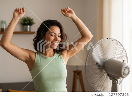Joyful Woman Celebrates Cool Relief Standing Near Electric Fan in Bright Modern Home Interior Joyful Woman Celebrates Cool Relief Standing Near Electric Fan in Bright Modern Home Interior 127797898