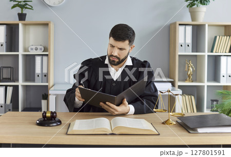 Man judge wearing judicial robe sitting at the desk with gavel and scales on workplace in courtroom 127801591