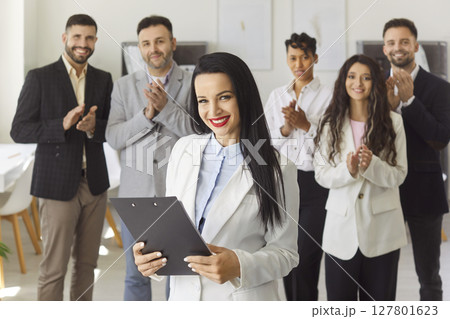Young woman looking at camera with team of company employees applauding in background in office. 127801623