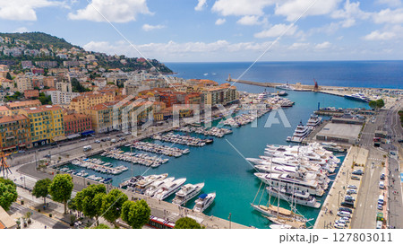 Drone view of the Port of Nice, France, showing luxury yachts, colorful historic buildings, blue Mediterranean Sea, and the coastline under a bright summer sky. 127803011