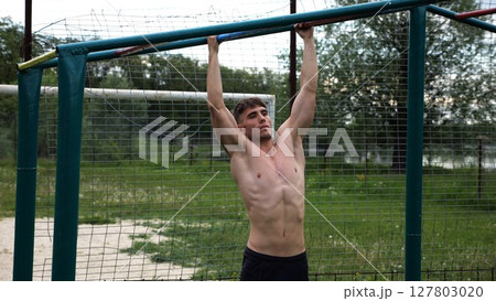 Young muscular sportsman doing pull ups at sports ground. Athletic guy exercising outdoor. Strong sporty man working out at summer nature. Concept of healthy and active lifestyle. Slow motion 127803020