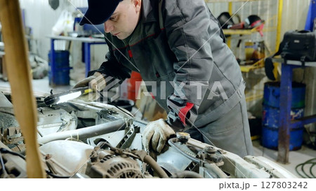 Skilled mechanic checking car engine with flashlight, wearing protective gloves during diagnostic evaluation in professional automotive workshop 127803242