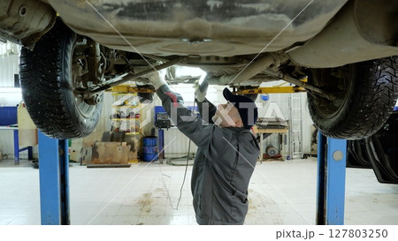 Experienced mechanic checking vehicle undercarriage with flashlight, working carefully in professional automotive repair workshop, wearing protective gloves Experienced mechanic checking vehicle undercarriage with flashlight, working carefully in professional automotive repair workshop, wearing protective gloves 127803250