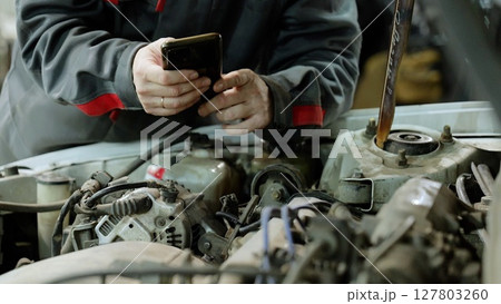 Car mechanic checking vehicle engine using smartphone, displaying digital diagnostic technology in automotive maintenance 127803260