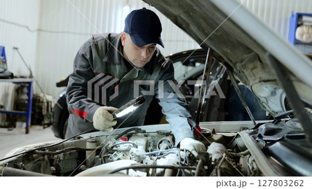 Professional mechanic inspecting vehicle engine, wearing protective gear and using flashlight while performing detailed diagnostic work inside modern automotive repair facility Professional mechanic inspecting vehicle engine, wearing protective gear and using flashlight while performing detailed diagnostic work inside modern automotive repair facility 127803262