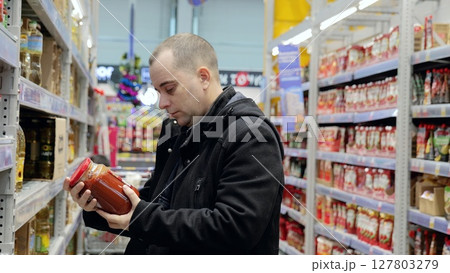 Male shopper selecting tomato sauce jar, examining product label, comparing brands while grocery shopping in supermarket aisle Male shopper selecting tomato sauce jar, examining product label, comparing brands while grocery shopping in supermarket aisle 127803279