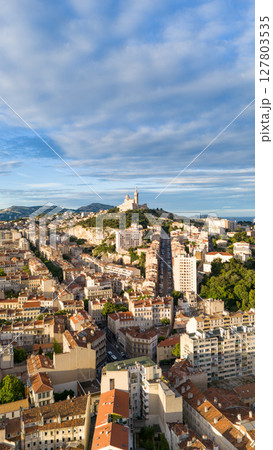 Drone aerial view of the city of Marseille, France, with the iconic Notre-Dame de la Garde basilica on a hilltop surrounded by urban buildings under a partly cloudy sky. Drone aerial view of the city of Marseille, France, with the iconic Notre-Dame de la Garde basilica on a hilltop surrounded by urban buildings under a partly cloudy sky. 127803535