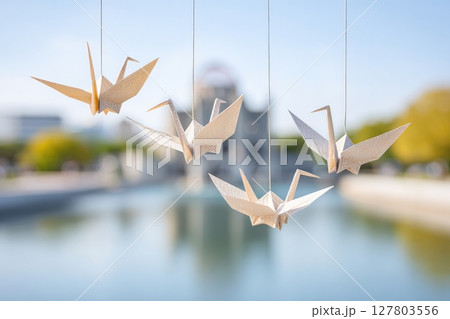 Delicate paper cranes are hanging above a calm water surface, surrounded by vibrant greenery, embodying themes of peace and tranquility in a serene environment 127803556