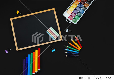 black chalk board and school chalk, felt-tip pens, paints, pen, paper clips, rubber band, on black background top view 127804672