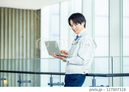 オフィスの廊下でノートパソコンを持つ作業服の若い作業員 オフィスの廊下でノートパソコンを持つ作業服の若い作業員 127807328