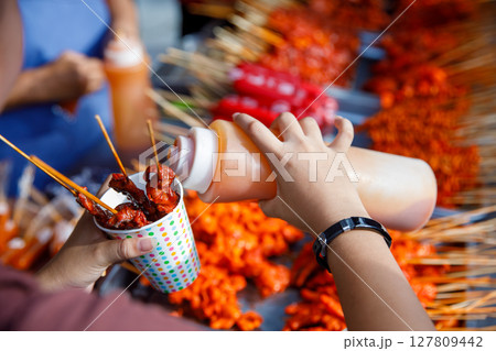 Filipino street food skewers with red sauce in paper cup, hand pouring sauce at outdoor market 127809442