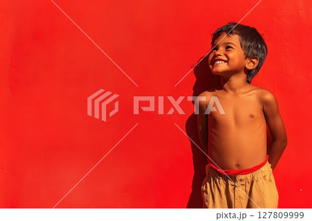 Young boy with playful grin against bright red background in sunlight 127809999