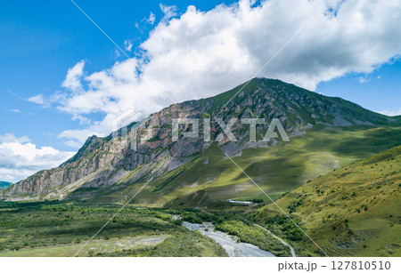 Majestic mountains rise dramatically against a bright blue sky, surrounded by verdant valleys and gentle slopes Majestic mountains rise dramatically against a bright blue sky, surrounded by verdant valleys and gentle slopes 127810510