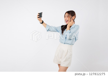 Happy young excited Asian woman laughing, taking a selfie picture with her smart mobile phone. She is making a quick selfie for social media. make video call isolated studio white background 127812275
