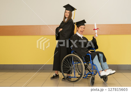 Happy young woman and woman in wheelchair in graduate gown with diploma in hands. Inclusive education. Happy young woman and woman in wheelchair in graduate gown with diploma in hands. Inclusive education. 127815603