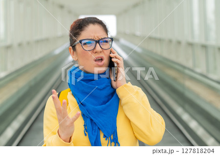 Caucasian woman stands on the escalator and is displeased talking on the mobile phone. 127818204