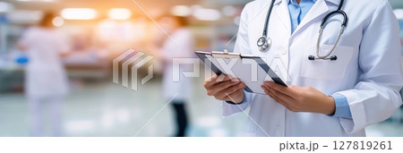 Doctor in white coat and stethoscope reviewing medical records in a busy hospital with blurred medical personnel, highlighting healthcare professionalism and patient care 127819261