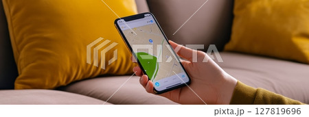 Woman is holding smartphone displaying online map with marked location, using mobile navigation app for route planning and location search while sitting on comfortable sofa with yellow pillows 127819696